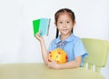 Asian little girl in school uniform sitting at table showing account book and carry piggy bank isolated over white background. Royalty Free Stock Photo