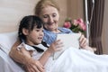 Lovely girl and grandmother watching fun and happy video together on tablet on patient bed. granddaughter visit the sick Royalty Free Stock Photo