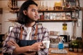 Asian male barista stands at a casual cafe, looking outside with a coffee cup