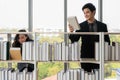 Asian male and cuacasian female university students standing at bookshelf seaching and reading book in library