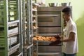 Asian male pastry chef taking stock of sweet bread from bread rack inside indoors kitchen with big oven