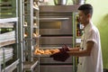 Asian male pastry chef taking stock of sweet bread from bread rack inside indoors kitchen with big oven