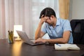 Asian man having severe migraine headache due to long hours of work and reserving a laptop at his desk at home