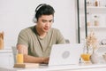 Asian man in headphones using laptop while sitting at kitchen table Royalty Free Stock Photo