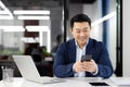 Asian man inside office with phone in hands, smiling contentedly, businessman using smartphone sitting at desk, mature Royalty Free Stock Photo