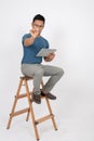 An Asian man is pointing his pen at the camera, holding his digital tablet, and sitting on a ladder Royalty Free Stock Photo