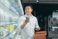 asian man shopper walks around the supermarket with a shopping cart looking at smart phone browsing smartphone in hands Royalty Free Stock Photo