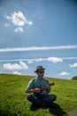 An Asian man sitting on a hill at a park, strumming a ukulele.