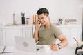 Asian man talking on smartphone while sitting at kitchen table near laptop Royalty Free Stock Photo