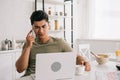 Asian man talking on smartphone while sitting at kitchen table and using laptop Royalty Free Stock Photo