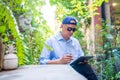 Asian man wearing a blue shirt, hat and sunglasses, sitting and working with a tablet in the garden Royalty Free Stock Photo