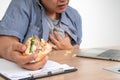 Asian man working and eating a burger on the office desk and heart attack. Concept of a busy businessman cannot work-left balance Royalty Free Stock Photo