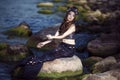 Asian Mermaid Posing Near Ocean Shore on Rocks While Wearing Seashell Decorated Crown and Black Shiny Tail On Seductive Body