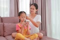 Asian mother braided hair for her little girl child while playing smartphone on sofa in living room at home Royalty Free Stock Photo