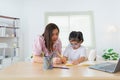 Asian mother teaching baby girl wearing eyeglasses doing home using laptop and write notes in notebook to study online on wood Royalty Free Stock Photo