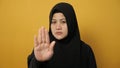 Asian muslim lady wearing hijab shows stop sign, prohibit forbid disallow something bad with palm hand gesture, against yellow