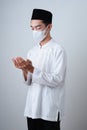Asian Muslim Man wearing muslim clothes with medical mask and skull cap praying to God against on grey Royalty Free Stock Photo