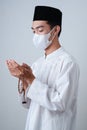 Asian Muslim Man wearing muslim clothes holding prayer beads with medical mask and skull cap praying to God against on grey Royalty Free Stock Photo