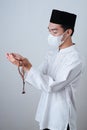 Asian Muslim Man wearing muslim clothes holding prayer beads with medical mask and skull cap praying to God against on grey Royalty Free Stock Photo