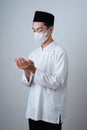 Asian Muslim Man wearing muslim clothes with medical mask and skull cap praying to God against on grey Royalty Free Stock Photo