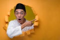 Asian Muslim Teenager man poses through torn yellow paper hole with shocked face, wearing muslim cloth with skull cap, pointing a Royalty Free Stock Photo