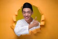 Asian Muslim Teenager man smiling at the camera with thumb up to copy sapce, poses through torn yellow paper hole, wearing muslim Royalty Free Stock Photo