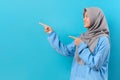Asian muslim woman points away on copy space, with hijab, feels optimistic, isolated over blue background