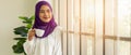 Asian muslim woman standing by window and having coffee or tea