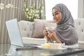 Asian Muslim woman working in living room, holding a coffee cup and using a laptop computer Royalty Free Stock Photo