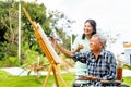 Asian nurse woman stand in the back to cheer up senior man on wheelchair to paint the painting outdoor and they look happy Royalty Free Stock Photo