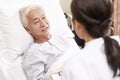 Asian old man lying in bed talking to young female doctor in hospital ward Royalty Free Stock Photo
