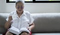 Asian older woman holding cup of coffee and reading a book beside the window after get up in morning Royalty Free Stock Photo