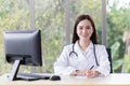 Asian professional female doctor who wears medical coat is working in office room at hospital while has computer put on table Royalty Free Stock Photo