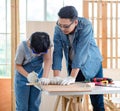 Asian professional male carpenter woodworker engineer dad in jeans outfit with safety gloves goggles helping teaching young boy Royalty Free Stock Photo