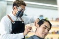 Asian professional male hairstylist combing and use scissors cutting young man customer`s hair in salon. Hairdresser wearing prote Royalty Free Stock Photo
