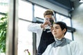 Asian professional male hairstylist combing and use scissors cutting young man customer`s hair in salon. Hairdresser wearing prote Royalty Free Stock Photo