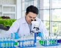 Asian professional male scientist researcher in white lab coat and rubber gloves sitting using microscope inspecting quality of Royalty Free Stock Photo