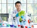 Asian professional male scientist researcher in white lab coat rubber gloves sitting using microscope looking at green salad Royalty Free Stock Photo