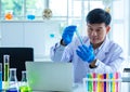 Asian professional mature male scientist in white lab coat and rubber gloves sitting holding looking at blue liquid sample in test Royalty Free Stock Photo