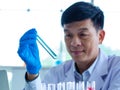 Asian professional mature male scientist in white lab coat and rubber gloves sitting holding looking at blue liquid sample in test Royalty Free Stock Photo