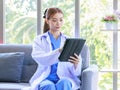 Asian professional successful female doctor in white lab coat with stethoscope sitting smiling holding tablet computer posing in Royalty Free Stock Photo