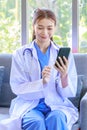 Asian professional successful female doctor in white lab coat with stethoscope sitting smiling using mobile phone posing in Royalty Free Stock Photo
