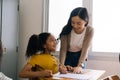 Asian school teacher assisting female student in classroom. Young woman working in school helping girl with her writing Royalty Free Stock Photo