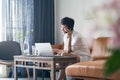 Asian self employed woman talking on smartphone on couch & working on laptop at home Royalty Free Stock Photo