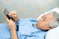 Asian senior or elderly old lady woman patient holding in her hands digital tablet and reading emails while sitting on bed in Royalty Free Stock Photo