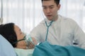 Asian senior female patient with oxygen mask lying on bed in hospital with asian male doctor examining patient with stethoscope