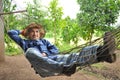 An asian senior gardener in blue checkered shirt,wears hat, lying on a hammock while resting in his garden after working Royalty Free Stock Photo