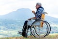 Asian senior man hold a cup of tea and sit on wheelchair with lood forward and stay near the cliff and mountain and grassfield as Royalty Free Stock Photo