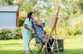 Asian senior man lean and relax action on wheelchair also look to painting after finish and nurse stay beside in the garden Royalty Free Stock Photo