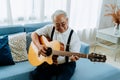 Asian senior man sitting on the sofa and playing acoustic guitar. Happy smiling elderly singing and enjoying with guitar Royalty Free Stock Photo